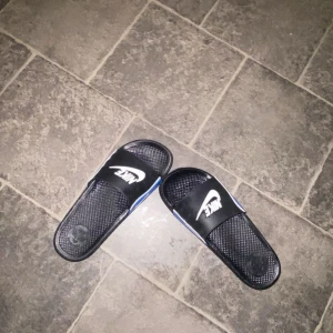 Svarta och blå Nike sandaler - Säljer ett par svarta och blå Nike sandaler med vit logga. Perfekta för avslappnade dagar och sommarbruk. Köptes för 200 kr  den funkar för tjejer och killar att ha den men de passar mer på killar/man