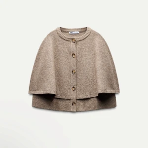 Beige cape från Zara - Snygg beige cape från Zara med knappar framtill. Perfekt för lager-på-lager under höst och vår. Den har en rund hals och ribbade kanter som ger en stilren look.