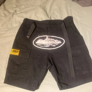 Svarta shorts från Corteiz - Snygga svarta shorts från Corteiz med coolt tryck och praktiska fickor. De har en justerbar rem i midjan och flera fickor med detaljerade broderier. Perfekta för en avslappnad stil.