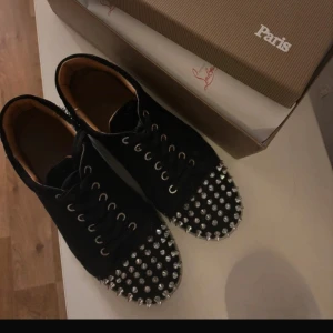 Svarta sneakers med nitar från Christian Louboutin - Snygga svarta sneakers från Christian Louboutin med silverfärgade nitar och den ikoniska röda sulan. Skorna har snörning och en unik design som verkligen sticker ut. 4 nitar är av men det är inget som märks, box och allt tillkommer såklart🖤