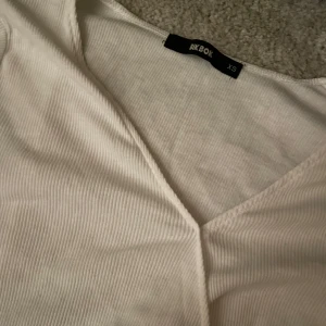 Vit ribbad omlottopp från Bik Bok - Säljer en stilren vit ribbad omlottopp från Bik Bok i storlek XS. Toppen har en v-ringad design och långa ärmar, perfekt för en chic och bekväm look.i bra skick 