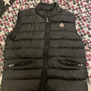 Svart dunväst från Moncler - Snygg svart dunväst från Moncler med dragkedja och hög krage. Västen har två praktiska fickor med dragkedja framtill och Monclers logga på bröstet. Storlek L