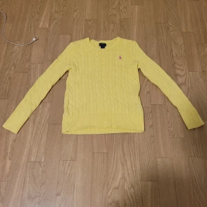 Gul stickad tröja från Ralph Lauren - Säljer en snygg gul stickad tröja från Ralph Lauren med kabelstickat mönster. Tröjan har långa ärmar och en rosa broderad logotyp på bröstet. Perfekt för att lysa upp garderoben!
