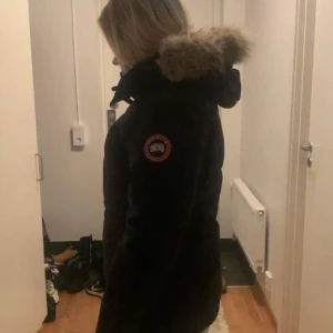 Svart parkajacka från Canada Goose - Säljer en svart parkajacka från Canada Goose med pälsdetaljer på huvan. Jackan har en klassisk design med lång ärm och är perfekt för kalla vinterdagar. Märkets logga syns tydligt på ärmen. Storlek M passar även s