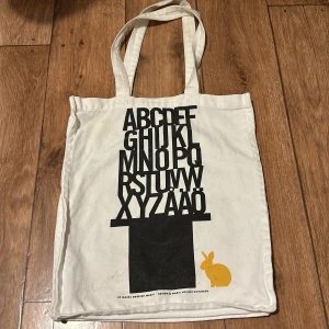 Vit shoppingväska med alfabetstryck - Säljer en vit shoppingväska från Ammunition Design med ett coolt alfabetstryck i svart och en gul kanin. Perfekt för att bära med sig böcker eller andra småsaker. Väskan har långa handtag för enkel bärning.