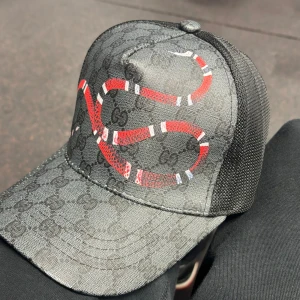 Svart keps från Gucci med ormmönster - Snygg svart keps från Gucci med ett iögonfallande ormmönster i rött och vitt. Kepsen har ett justerbart band baktill med blå och röda detaljer. Perfekt för att ge din outfit en lyxig touch.