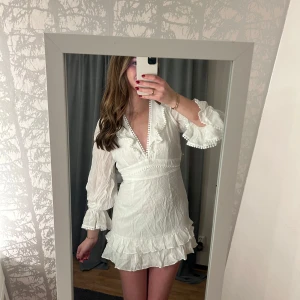 Vit studentklänning showpo - Charmig vit klänning med volanger och snörning i ryggen. Den har en v-ringad design med dekorativa detaljer och långa ärmar. Perfekta studentklänningen!💕Nypris $79.95