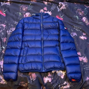 Blå dunjacka från Tommy Hilfiger - Säljer en snygg blå dunjacka från Tommy Hilfiger. Jackan har en quiltad design med dragkedja och långa ärmar. Den är perfekt för kyligare väder och har en stilren look med märkets logga på bröstet. Äkta såklart passar barn från 10-14 väldigt skön. Säljer den då den är för liten