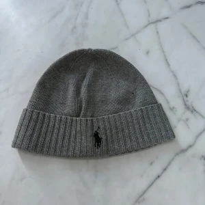 Grå stickad mössa från Ralph Lauren - Snygg grå stickad mössa från Ralph Lauren med broderad logga framtill. Perfekt för kyliga dagar och en stilren look.