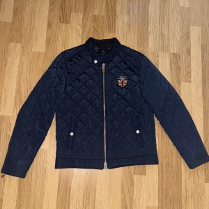 Mörkblå quiltad jacka från Morris - Snygg mörkblå quiltad jacka från Morris med dragkedja och broderad logotyp på bröstet. Jackan har en stilren design med långa ärmar och knappdetaljer vid kragen. Perfekt för en stilmedveten look.