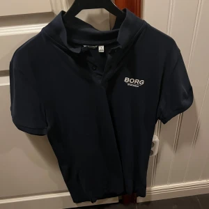 Mörkblå pikétröja från Björn Borg - Snygg mörkblå pikétröja från Björn Borg med korta ärmar och klassisk krage. Perfekt för en sportig look eller avslappnad stil. Diskret logga på bröstet.