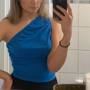 Blå oneshoulder topp - Snygg blå oneshoulder topp med en elegant drapering över axeln. Perfekt för en kväll ute eller en speciell tillställning. Toppen har en tight passform som framhäver figuren.