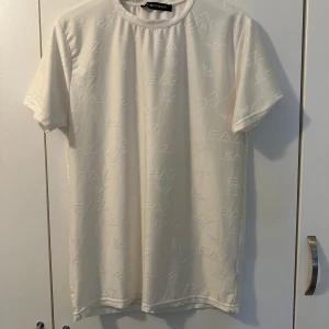 Vit t-shirt från Emporio Armani - Snygg vit t-shirt från Emporio Armani med diskret EA7-logga över hela tyget. Perfekt för en stilren look. T-shirten har en normal passform och är kortärmad.