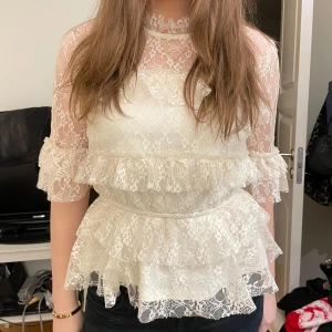 Vit spetsblus med volanger - Elegant vit blus i spets med volanger och trekvartsärm. Perfekt för en stilfull look.