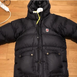 jacka från Fjällräven - Snygg och varm svart jacka från Fjällräven med huva och gula detaljer. Jackan har en dragkedja och knappar framtill samt ett emblem på bröstet. Perfekt för kyliga dagar.