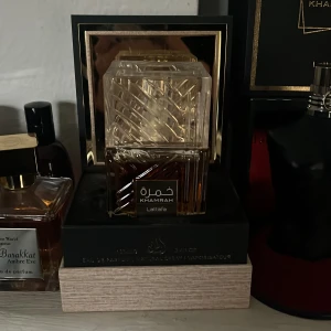 Khamrah Eau de Parfum från Lattafa - Elegant parfymflaska med ett unikt mönster och en klar, fyrkantig design. Khamrah från Lattafa erbjuder en lyxig doftupplevelse. Flaskan har en genomskinlig kork och en gyllene etikett som ger en exklusiv känsla. Dofter som en fin äppelpaj blandat med kanel! Knappt använd då den inte passar mig!