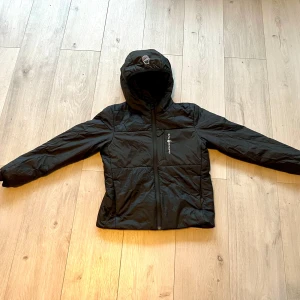 Svart jacka från Sail Racing stl 150 - Snygg svart jacka från Sail Racing med huva och dragkedja. Tillverkad med Primaloft-teknologi för extra värme och komfort. Jackan är vindtät och andas bra, perfekt för kyligare dagar. Storlek 150.