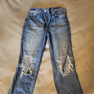 Blå jeans med slitningar från NA-KD - Säljer ett par coola blå jeans från NA-KD med slitningar på knäna. De har en klassisk femficksdesign och knappgylf. Perfekta för en avslappnad stil.