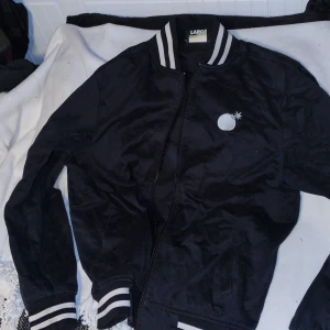 Svart college bomberjacka från The Hundreds - Snygg svart bomberjacka från The Hundreds med vit broderad logga på bröstet och ryggen. Jackan har dragkedja och ribbade muddar i svart och vitt vid ärmslut och nederkant. Perfekt för en cool och avslappnad stil.