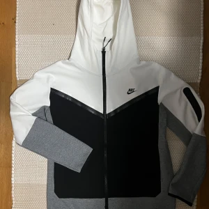 Svart och vit hoodie från Nike - Snygg hoodie från Nike i svart, vit och grå färgkombination. Den har en dragkedja framtill och en justerbar huva. Perfekt för en sportig look. Nike thec i storlek M, denna är den nya versionen, nike thec new season. Säljes för 699kr, knappt använt, nyskick
