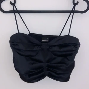 Svart satinlinnetopp från Gina Tricot - Snygg svart satinlinnetopp från Gina Tricot med smala axelband och rynkdetaljer framtill. Perfekt för en utekväll eller fest! Toppen är i bra skick och ger en elegant look.