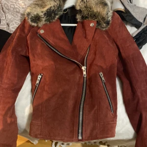 Röd mockajacka med pälskrage från Joeama - Snygg röd mockajacka med en lyxig pälskrage. Jackan har en asymmetrisk dragkedja och två fickor med dragkedjor. Perfekt för hösten och våren, ger en cool och stilren look. Passar både till vardags och fest!