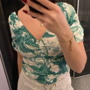 Grön och vit blommig blus - Säljer en superfin blus med grönt blommönster på vit bakgrund. Den har en v-ringad design och korta ärmar, perfekt för sommardagar. Blusen är i bra skick och passar både till vardags och festligare tillfällen (prislapp kvar). 