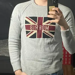 Säljer en snygg grå stickad tröja från Morris med Union Jack-motiv och texten 'Morris' på framsidan. Tröjan har långa ärmar och broderad detalj med 'KG 1968' på ärmen. Perfekt för höst och vinter! Bara att skriva vid frågor 😃