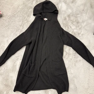 Svart stickad kofta med huva - Mysig svart stickad kofta med huva och långa ärmar. Perfekt för kyliga dagar. Den har praktiska fickor framtill och en avslappnad passform. Perfekt för både vardag och mysiga hemmakvällar.