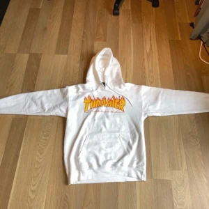 Vit hoodie från Thrasher - Säljer en snygg vit Thrasher hoodie med det klassiska logotyptrycket i gult och rött på bröstet. Den har en stor ficka fram och en bekväm huva. Perfekt för skate eller chill dagar. Passar till allt och är superbekväm!  Storlek S 34-36. Men funkar som XS. 90% bomull 10% polyester.
