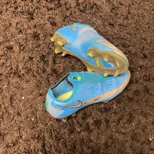 Blå Nike fotbollsskor - Säljer ett par blå Nike Phantom fotbollsskor i bra skick. De har en snygg design med silverdetaljer och är perfekta för fotbollsträning eller match. Skorna har en tight passform och är gjorda i syntetmaterial för bästa prestanda på planen. Dessa är nike Elite och kostar nypris 2799kr. Går lätt att svätta av.