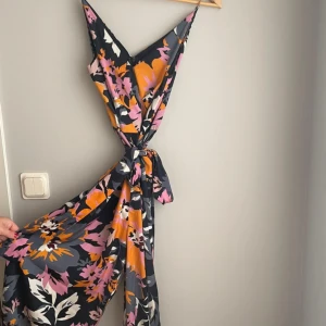 Blommig jumpsuit från H&M - Säljer en superfin blommig klänning från H&M i storlek 36. Den har ett färgglatt mönster i svart, rosa, orange och vitt. Klänningen är ärmlös och har en v-ringad design med knyt i midjan. Perfekt för sommarens fester eller en dag på stan!