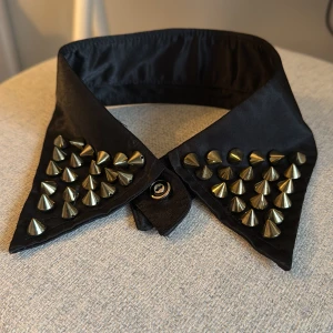 Svart krage med nitar - Snygg svart krage med coola guldfärgade nitar. Perfekt för att ge din outfit en edgy touch. Kragen har en knappstängning framtill och är lätt att matcha med olika stilar. Passar perfekt för festliga tillfällen eller när du vill sticka ut lite extra.