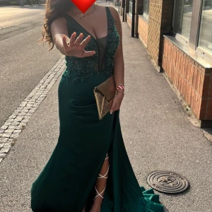 Grön långklänning med spetsdetaljer - Elegant grön långklänning med djup urringning och vackra spetsdetaljer. Klänningen har en hög slits och smala axelband, perfekt för festliga tillfällen. Den är figurnära och ger en glamorös look. Kombinera med en glittrig clutch för en komplett outfit. Den är använd en gång på en bal och är ren samt tvättat, den är köpt utomlands så ingen annan har en likadan!! Samt så har jag sytt om den och lagt till slits samt så har den dragkedja och band, där man kan välja vilken metod man vill ha vid rygge