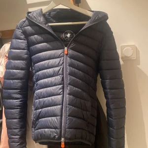 Mörkblå dunjacka från Save the Duck - Säljer en snygg mörkblå dunjacka från Save the Duck. Jackan är quiltad och har en dragkedja framtill. Perfekt för kyliga dagar med sin varma och lätta design. Den har en huva och elastiska muddar för extra komfort. Passar perfekt för höst och vinter! Priset går att diskutera