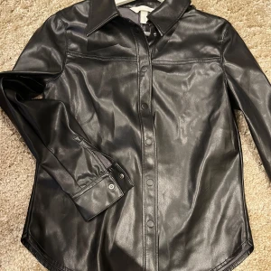 Svart skinnskjorta - Snygg svart skinnskjorta i mycket bra skick. Den har långa ärmar och knappar framtill. Perfekt för en cool och stilren look, oavsett om det är för en utekväll eller en avslappnad dag. Skjortan har en klassisk krage och är gjord av ett mjukt skinnmaterial som ger en lyxig känsla.