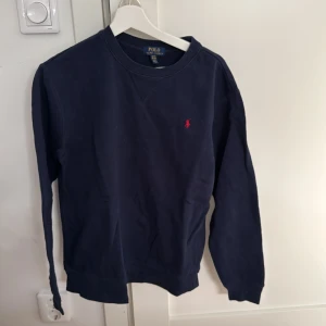 Mörkblå tröja från Polo Ralph Lauren - Säljer en snygg mörkblå tröja från Polo Ralph Lauren i storlek barn  XL. Tröjan har en klassisk design med ett rött broderat logotyp på bröstet. Perfekt för både vardag och lite finare tillfällen. Skön och stilren!