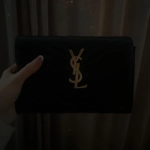 YSL väska  - Snygg svart kuvertväska med en elegant guldlogga på framsidan. Perfekt för festliga tillfällen eller en utekväll. Väskan har en stilren design och är lätt att matcha med olika outfits.