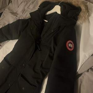 Säljer en svart parkajacka från Canada Goose i mycket bra skick. Jackan har en varm pälsfodrad huva och är perfekt för kalla vinterdagar. Den har en dragkedja framtill och flera praktiska fickor. Perfekt för dig som vill hålla dig varm och stilren i vinter! ❄️ nypris är 10000