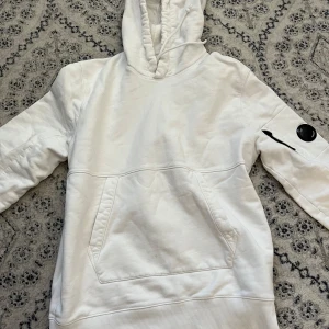 Vit hoodie från C.P. Company - Säljer en snygg vit hoodie från C.P. Company. Den har en klassisk design med en stor ficka fram och en cool detalj på ärmen. Perfekt för både vardag och chill. Hoodien är i bra skick och redo för nya äventyr!