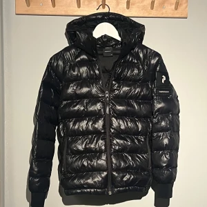 Svart glansig peak performance jacka - Säljer en otroligt snygg glansig svart pufferjacka från Peak Performance i barnstorlek 150. Jackan har en glansig finish och är perfekt för kyliga dagar. Den har en praktisk huva och dragkedja framtill. Perfekt för både vardag och utomhusaktiviteter! Öppen för byten.