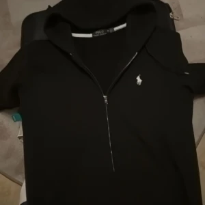 Svart tröja från Ralph Lauren - Säljer en stilren svart tröja från Ralph Lauren i storlek M. Tröjan har en dragkedja framtill och det klassiska logotypbroderiet på bröstet. Perfekt för både vardag och lite finare tillfällen. Passformen är custom fit vilket ger en snygg siluett.