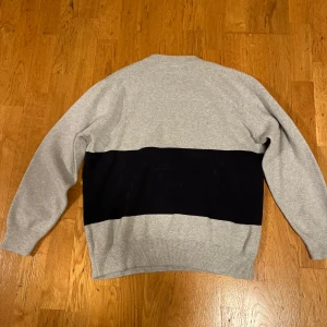 Grå och svart tröja från A Day's March - Säljer en stilren grå tröja med en bred svart rand från A Day's March. Tröjan är i medium storlek och har en klassisk passform med långa ärmar. Perfekt för både höst och vinter. Den är i mycket bra skick och redo för en ny ägare!