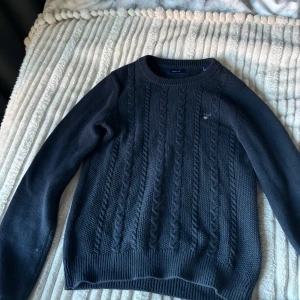 Mörkblå stickad tröja från GANT - Säljer en snygg mörkblå stickad tröja från GANT. Den har ett klassiskt flätmönster och en liten broderad logga på bröstet. Tröjan är långärmad och perfekt för kyliga dagar. Passar både till vardags och lite finare tillfällen. Supermysig och stilren!