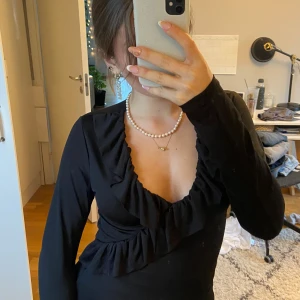 Svart långärmad topp med volangdetalj - Säljer en elegant svart topp med långa ärmar och en v-ringad design. Toppen har en snygg volangdetalj framtill som ger en extra touch. Perfekt för både vardag och fest!