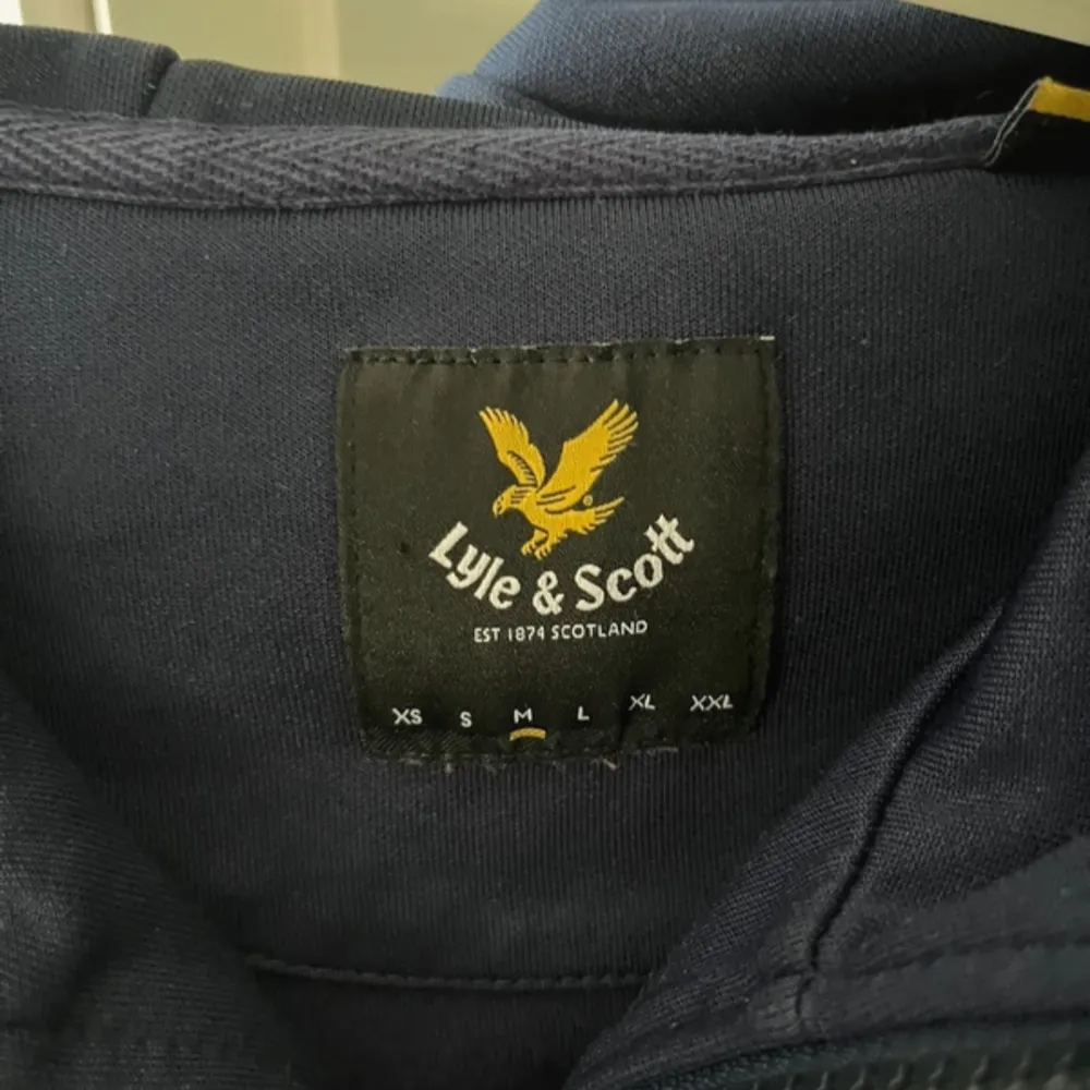 Säljer en snygg blå hoodie från Lyle & Scott i bra skick. Den har en dragkedja framtill och en gul broderad logga på bröstet. Perfekt för både höst och vår, med en skön passform och praktiska fickor. Perfekt för vardagsbruk eller en avslappnad dag ute.. Tröjor & Koftor.