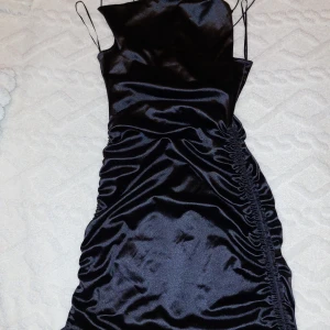 Svart satin klänning - Snygg svart klänning i satin med smala axelband och en elegant rynkad detalj på sidan. Perfekt för festliga tillfällen! Klänningen har en glansig finish som ger en lyxig känsla. Ny med taggar kvar.
