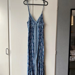 Blå långklänning med mönster - Säljer en superfin blå långklänning med ett snyggt mönster. Klänningen har smala axelband och en v-ringad design. Materialet känns lätt och luftigt.
