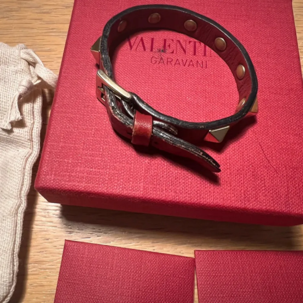 Snyggt vinrött läderarmband från Valentino med guldiga nitar. Armbandet har justerbar passform med flera hål och ett spänne i guld. Perfekt för att ge en edgy touch till din outfit. Kommer i originalförpackning med dustbag.. Accessoarer.