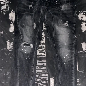 Svarta jeans från Dsquared2 - Säljer ett par svarta jeans från Dsquared2 i bra skick. De har en cool slitning och färgstänk som ger en edgy look. Jeansen är skinny fit och har knappar med Dsquared2-logga. Perfekta för en avslappnad men stilren outfit!